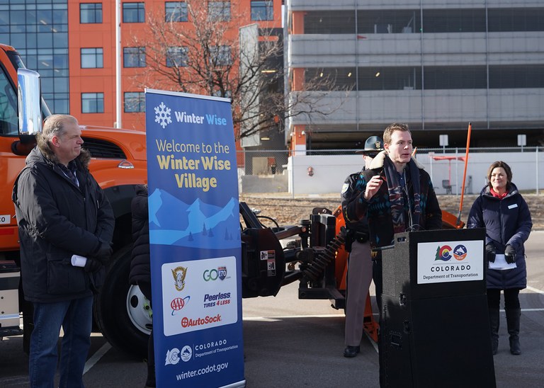 Members of the community look on as a man gives a speech in front of a CDOT snowplow