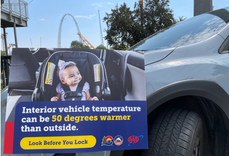 Look Before You Lock yard sign A yard sign rests against the front of a blue car. The sign has a baby in a car seat with the text overlay, “Interior vehicle temperature can be 50 degrees warmer than outside. Look before you lock.”