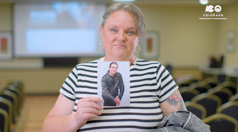 A woman standing in an empty conference room and holding up a photo of a young man. A woman standing in an empty conference room and holding up a photo of a young man. The CDOT logo is in the top right corner.