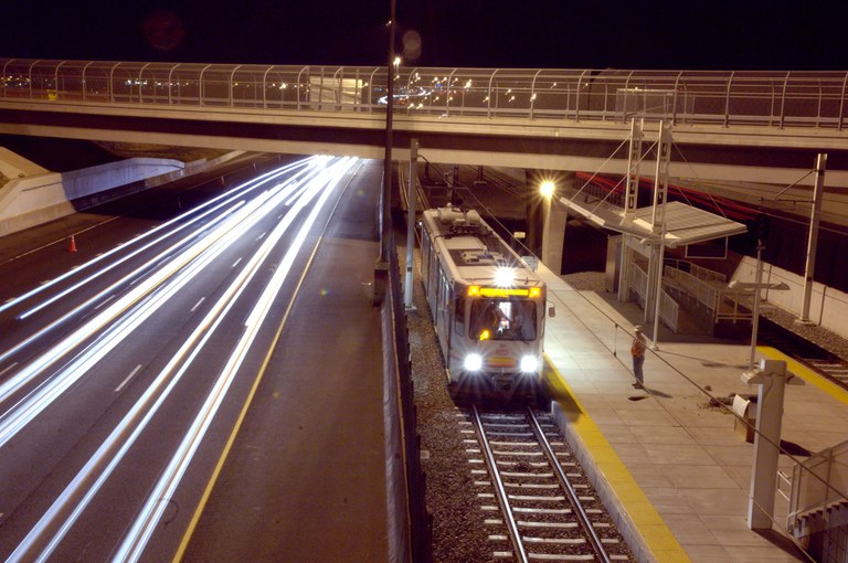 Nighttime shot of an RTD light rail train at an I-25 station platform alongside highway traffic light trails.