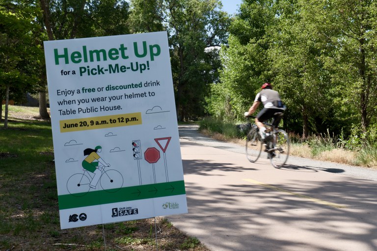 Sign reading, "Helmet Up for a Pick-Me-Up."  Enjoy a free or discounted drink when you wear your helmet to Table Public House, June 20, 9 a.m. to noon. The sign is placed next to a bike trail with a bicyclist in the background.  