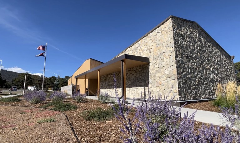 The fully renovated US 160 Shaw Creek Rest Area exterior. At the front of the building, an American flag and a Colorado state flag fly on a flagpole under a clear blue sky.