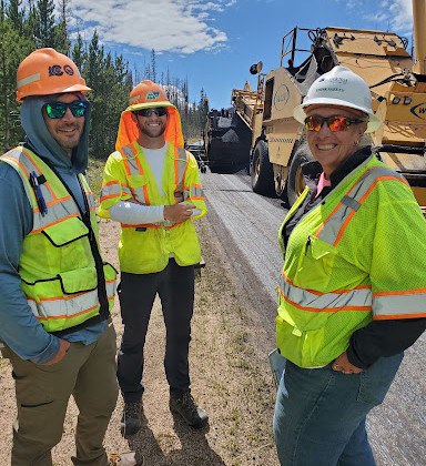 Three construction workers in high-visibility vests, hard hats, and sunglasses smiling next to paving equipment on a road job site.