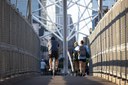 A bicyclist and several pedestrians with backpacks cross a modern suspension bridge with high metal mesh fencing on both sides. The view is from behind the travelers, looking through the white structural cables toward city buildings in the distance. thumbnail image