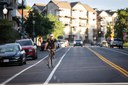 A person riding a bicycle down a dedicated white-striped bike lane on a multi-lane city street. Parked cars line the left side of the road, and residential apartment buildings are visible in the background under bright daylight. thumbnail image
