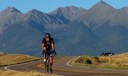 A bicyclist wearing a helmet and cycling jersey rides toward the camera on a paved two-lane road with a double yellow center line. The road curves through a grassy, open landscape. In the background, a massive range of jagged, blue-toned mountains rises against a clear sky. A car is parked on the shoulder in the distance on the right. Photo courtesy of Westcliffe Adventures' historic Bike with Pike, along the Frontier Pathways National Scenic and Historic Byway. thumbnail image