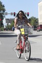 A smiling woman in sunglasses and a patterned shirt rides a red shared-use bicycle through a city intersection. The bike features a front basket with a yellow sign. In the background, city traffic lights and street signs are visible under a clear sky. thumbnail image