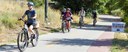 A group of five bicyclists wearing helmets ride along a paved multi-use path with a yellow dashed center line. A sign on the right side of the path features a black arrow and the text "Bike to Work Day Station." Dense green foliage and dry grass border the trail. thumbnail image