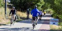 Several cyclists wearing helmets ride along a paved multi-use path with a yellow dashed center line. A sign on the right side of the trail features a black arrow and the text "Bike to Work Day Station." The trail is surrounded by green trees and dry grass. thumbnail image
