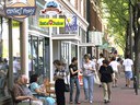A sidewalk scene in downtown Fort Collins with several pedestrians walking. Business signs featuring clear text are prominent. Signs visible from top left include: The Clothes Pony, Mary's Mountain cookies, Inta Juice, and Daisy Sports. The scene also shows a group of seniors sitting and talking on benches on the left side. Photo courtesy of Fort Collins. thumbnail image
