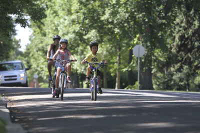 An adult woman and two young children ride bicycles toward the camera on a paved neighborhood road. All three are wearing helmets. The road is lined with lush green trees, and a white car is visible in the background on the left. A stop sign stands further back on the right side of the road. Photo courtesy of Bicycle Colorado.