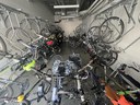 A narrow indoor bicycle parking room filled with many bikes. Rows of bicycles are hanging vertically from metal wall-mounted racks on both sides, while several other bicycles are parked on the floor in the center. The room has concrete floors and white walls. thumbnail image