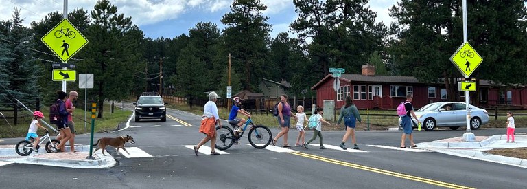 Pedestrian crossing near Buffalo Park A group of pedestrians, children on bicycles, and a dog walking across a marked crosswalk on a paved road flanked by yellow pedestrian crossing signs, with cars stopped nearby and pine trees in the background.
