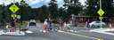 A group of pedestrians, children on bicycles, and a dog walking across a marked crosswalk on a paved road flanked by yellow pedestrian crossing signs, with cars stopped nearby and pine trees in the background. thumbnail image