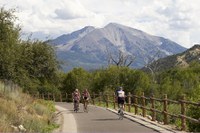 Four cyclists riding on the paved Rio Grande Trail. Three cyclists are riding toward the camera, and one is riding away. The trail is bordered by a rustic wooden post-and-rail fence and lush green trees. In the background, a large, prominent mountain peak stands under a sky filled with large, white, puffy clouds. Photo courtesy of VisitGlenwood.com
