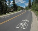 A paved two-lane road with a double yellow center line curves through a forest. Large white shared lane markings, or "sharrows," consisting of a bicycle symbol and two chevrons, are painted in the center of the travel lanes. Pine trees and a log building are visible on the sides of the road under a partly cloudy sky. Photo courtesy of the Town of Breckenridge thumbnail image