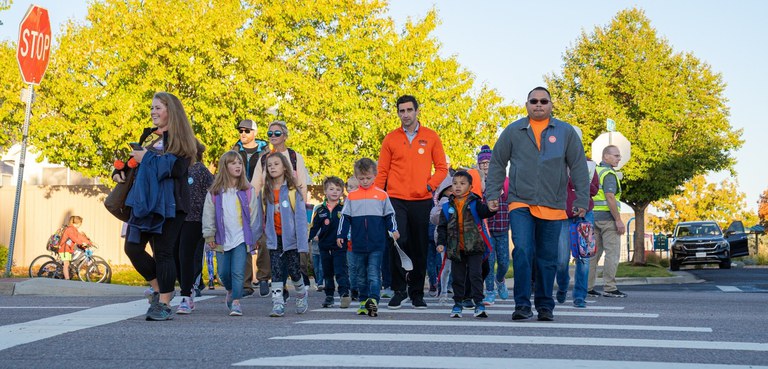 A group of elementary school children and adults walking together across a painted street crosswalk on a sunny day with bright autumn trees and a stop sign in the background. Photo courtesy of Canyon Creek School District. 
