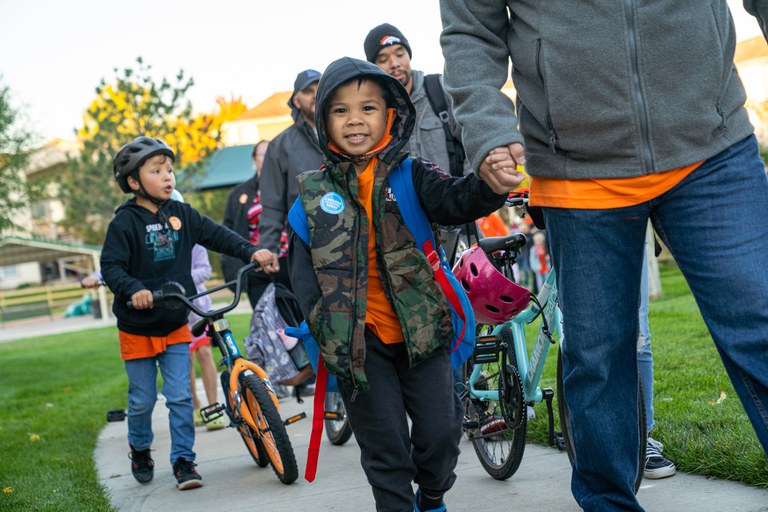 A smiling young boy wearing a hooded vest holding an adult's hand as they walk along a sidewalk on Walk to School Day, accompanied by other students and parents riding bicycles. Photo courtesy of Canyon Creek School District.