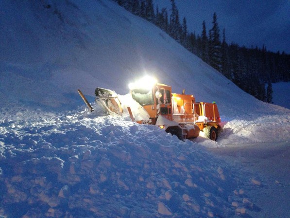 An orange, CDOT snowplow with its headlight on clears snow at night.