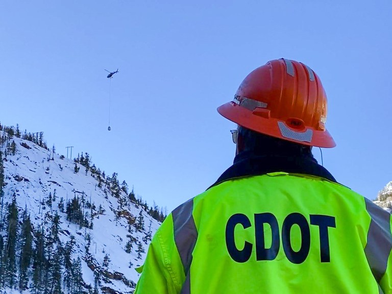 A member of CDOT's specially trained avalanche crew observes avalanche mitigation equipment being transported into place by a helicopte