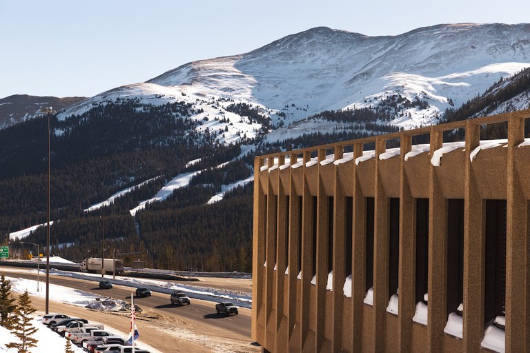 A highway with light traffic running past a concrete tunnel structure, set against a backdrop of snow-capped mountains and evergreen forests.