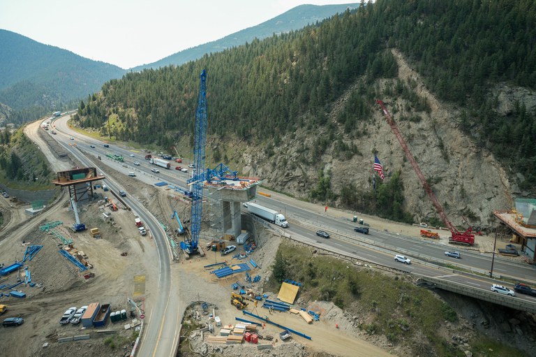 Aerial view of interstate tucked between mountains with construction activity. A large bridge pier is being constructed with a blue crane nearby.