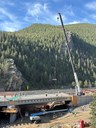 A high view of a bridge being built in front of a giant mountain covered in pine trees. A tall crane stands on the right, lowering a piece of equipment down to workers on the new bridge deck. thumbnail image