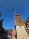 A low-angle shot of a tall concrete bridge pier under construction in a narrow mountain canyon. A large red lattice boom crane towers over the structure. thumbnail image