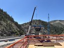 A large concrete bridge pier under construction in a mountain canyon. A white mobile crane and a tall blue lattice boom crane stand next to the pier. An interstate highway runs along the canyon floor to the left. thumbnail image