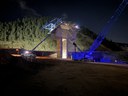A low-angle night view of a bridge construction site in the mountains. Bright work lights light up a concrete support pillar with scaffolding on its side. A large blue crane stands next to it, and a pickup truck with flashing lights sits nearby. thumbnail image