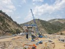 A high-angle view of a bridge construction project in a mountain canyon. A concrete support pier stands centrally, topped with a blue steel framework. A multi-lane highway wraps around the canyon hillside with semi-trucks and traffic passing by. thumbnail image