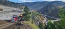 An elevated view of a major highway construction project in a mountain canyon. A red semi-truck drives on a dirt road next to a large concrete retaining wall and a yellow excavator. Down in the valley, yellow drilling rigs and construction vehicles operate alongside a highway winding through the hills. thumbnail image