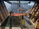 Construction crew performing a concrete bridge deck pour at sunrise with a concrete pump arm and crane silhouetted against a vibrant orange mountain sky. 