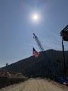 A large American flag hangs from a blue construction crane along a dirt access road, backlit by a bright sun over a pine-covered mountain ridge. thumbnail image
