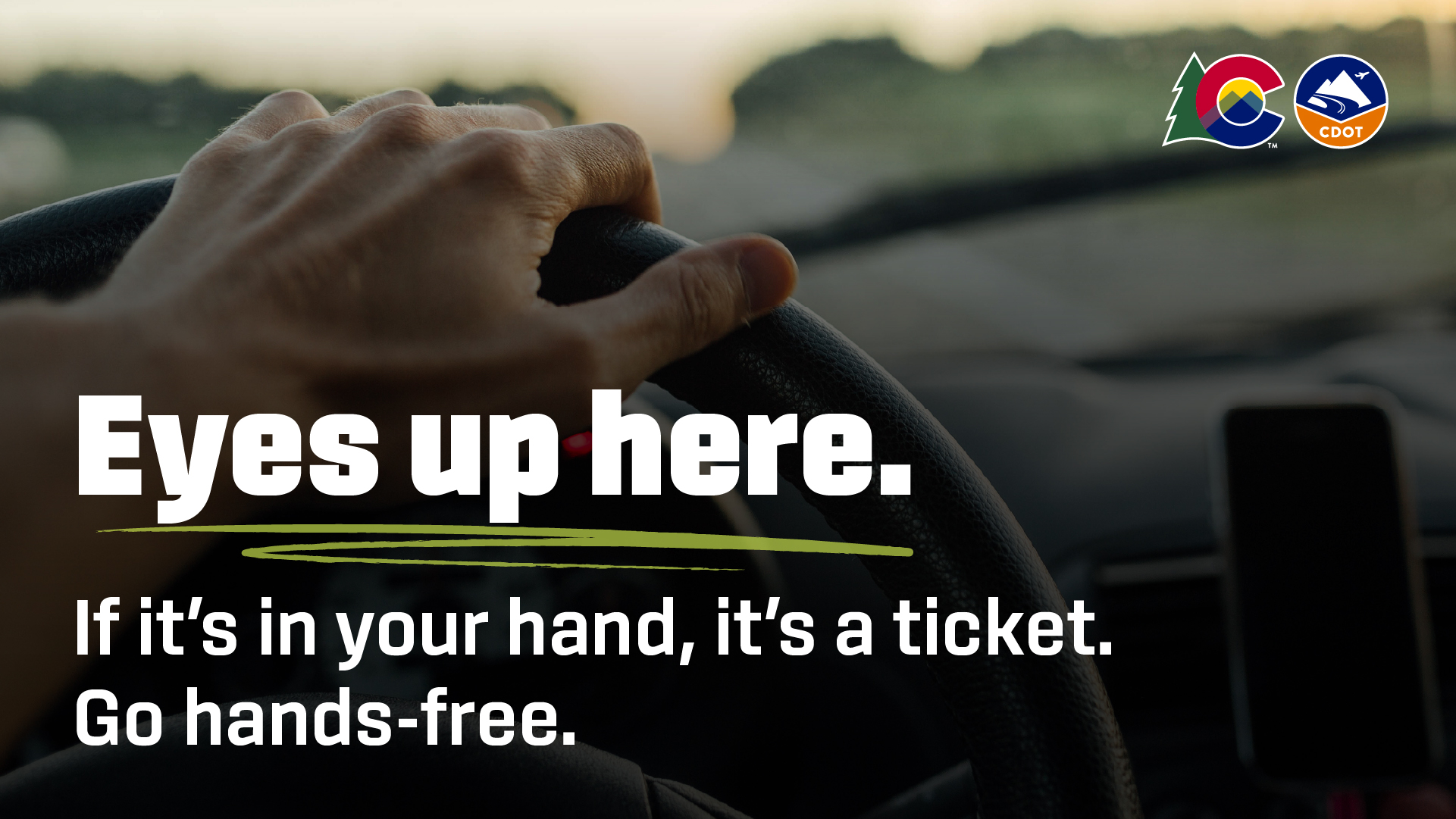 A hand on steering wheel with text overlay reading "eyes up here. If it's in your hand, it's a ticket. Go hands free"