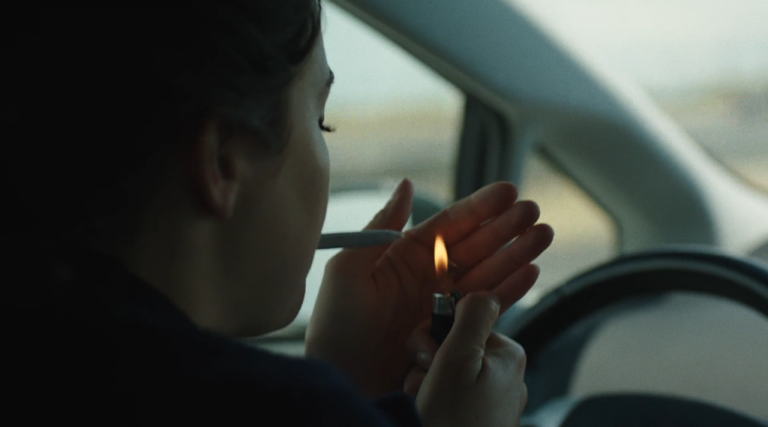 A woman driver holds both hands off the steering wheel to light a cannabis joint.