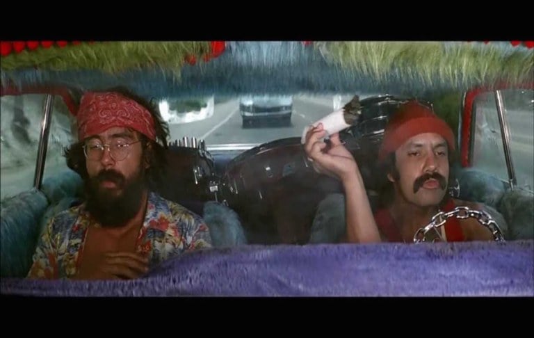 Two people in a car wearing colorful clothing. The driver is holding a large cannabis joint. 