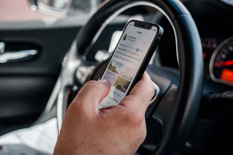 Persons hand shown scrolling through social media on cell phone behind a steering wheel