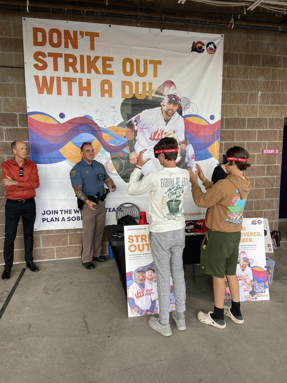 Teen boys try to catch a ball while wearing impaired simulation goggles in front of a banner reading “Don’t Strike Out with a DUI.”