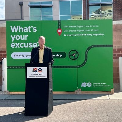 A news conference takes place with CDOT Traffic Safety Communications Manager Sam Cole speaking at a podium in a paved lot. Behind him, a large green CDOT display billboard that says, "Most crashes happen close to home. Most crashes happen under 40 miles per hour. So wear your seat belt every single time."