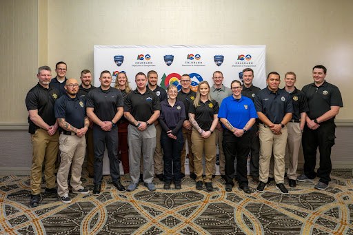 A group of Drug Recognition Expert (DRE) graduates smiling standing in front a a backdrop with the Highway Safety Office Occupant Protection Assessment logo and the Colorado Department of Transportation logo on it. 