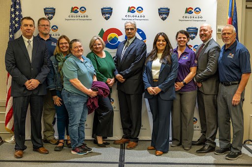 A group of Law Enforcement Liaisons smiling standing in front a a backdrop with the Highway Safety Office Occupant Protection Assessment logo and the Colorado Department of Transportation logo on it. The Colorado state flag and American flag are on either side of the backdrop. 