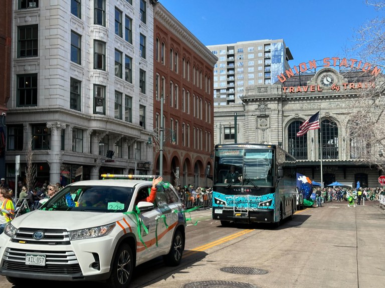 A CDOT vehicle bedecked in St. Paddy's Day decorations driving down downtown Denver followed by a Bustang Outrider bus. Union Station is in the background as people cheer from the sidewalks.