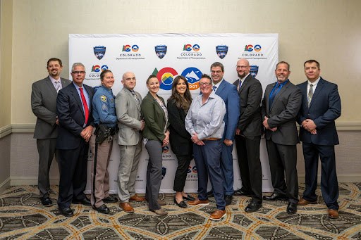 A group of Drug Recognition Expert (DRE) graduates smiling standing in front a a backdrop with the Highway Safety Office Occupant Protection Assessment logo and the Colorado Department of Transportation logo on it. 