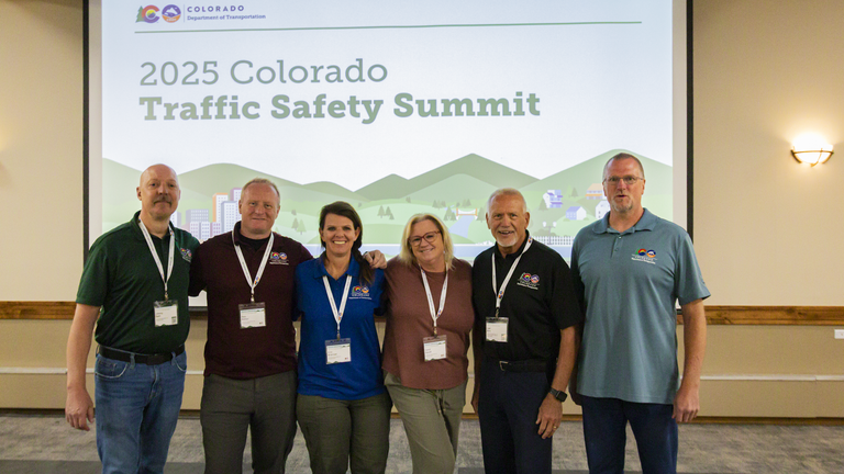 Traffic Safety Summit attendees in colorful polos stand in front of a screen with an illustrated image of green mountains and blue sky with an array of transportation modes and city buildings. Text overlay reads, "2025 Colorado Traffic Safety Summit." 
