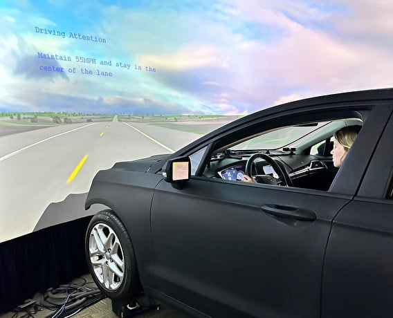 Human behavior insights are driving transportation safety forward Person sitting in the driver’s seat of a black vehicle while using an immersive driving simulator that’s displaying visuals of a roadway and blue sky with pink clouds.