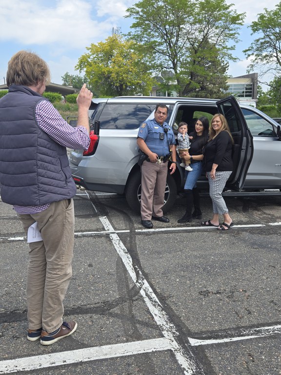 Colorado State Patrol trooper, women and child posing in front of a grey SUV as a cameraman films them in a parking lot. 