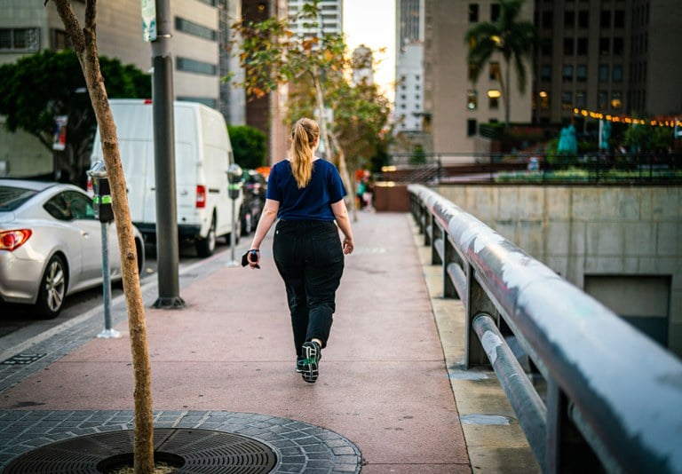 Woman walking on city sidewalk with cars parked on the street and tall buildings and trees lining the road.