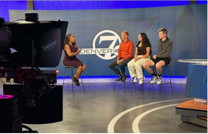 A television studio interview showing four people seated in front of a backdrop with the Denver 7 logo. A female interviewer holds a microphone while speaking with three guests – a man in an orange/red shirt, a young woman with long dark hair in a black top and light-colored pants, and a young man in a gray hoodie. Professional television cameras and equipment are visible in the foreground, with blue accent lighting and a curved studio floor design typical of a news broadcast setting.