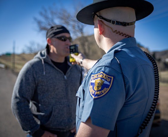 Colorado State Patrol trooper in blue uniform administering a roadside breathalyzer test to a man in a grey jacket and black hat.